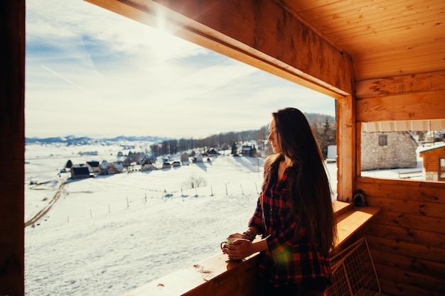 Młoda kobieta trzymająca kubek, stojąca przy oknie w domku w zimie