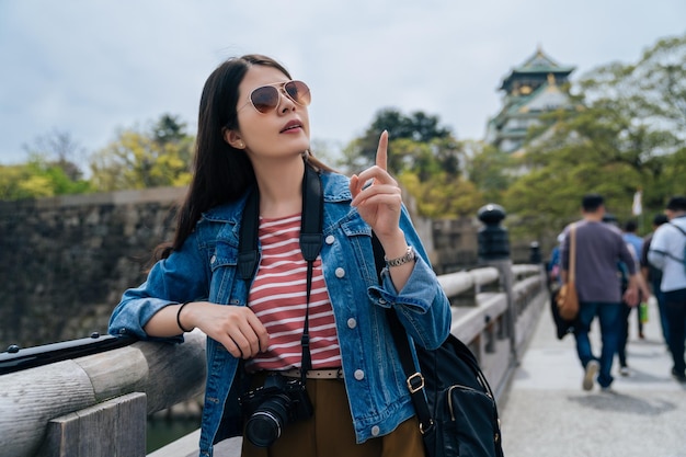 Zdjęcie młoda kobieta turysta z okularami przeciwsłonecznymi odwiedza zamek w japonii i relaksuje palcem wskazującym na błękitne niebo. wiele osób idzie do wieży osaka. kobieta obiektyw mężczyzna stojący opierając się na poręczy prowadzenia aparatu.