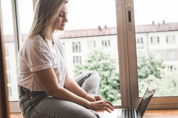 Młoda Kobieta Ubrana Od Niechcenia, Pracując Na Laptopie, Siedząc Na Parapecie W Domu. Praca W Domu W Koncepcji Przytulnej Atmosfery