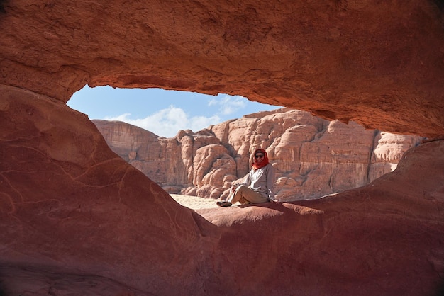 Młoda kobieta ubrana w ciepłą kurtkę i szalik wokół głowy uśmiechnięta, siedząca przy naturalnie uformowanym skalnym oknie na pustyni wadi rum w słoneczny dzień