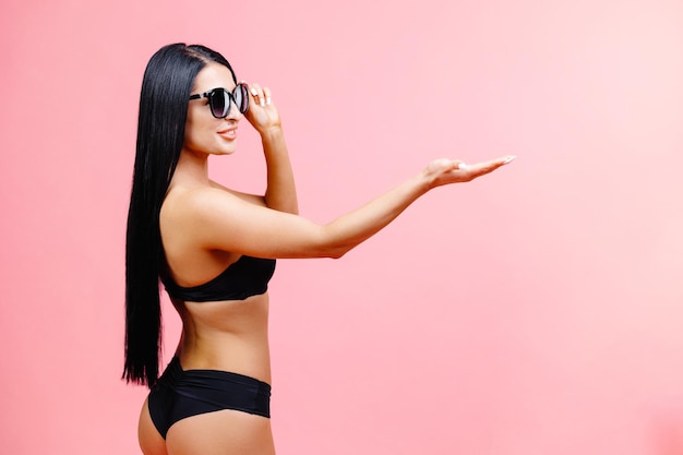 Młoda kobieta ubrana w czarny strój kąpielowy i okulary przeciwsłoneczne wskazując ręką na obszar promocyjny makiety kopii przestrzeni na białym tle na różowym tle studio