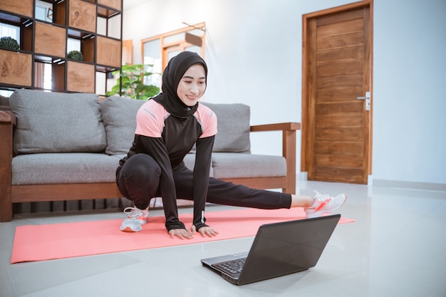 Młoda kobieta ubrana w hidżab w stroju gimnastycznym podczas kucania rozciąga się z jedną nogą wyciągniętą na bok przed laptopem w domu
