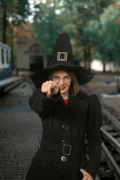 Młoda kobieta ubrana w kapelusz czarownicy czarny płaszcz i okulary z magiczną różdżką w rękach Uśmiechnięta dziewczyna Pionowa ramka