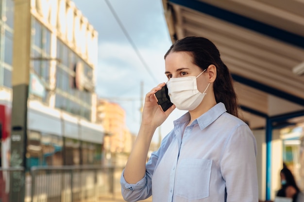 Młoda kobieta ubrana w maskę i rozmawia przez telefon, czekając na transport publiczny.