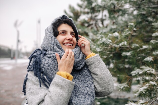 Młoda kobieta ubrana w szary płaszcz i niebieski szalik rozmawia przez telefon komórkowy w snowy winter park.