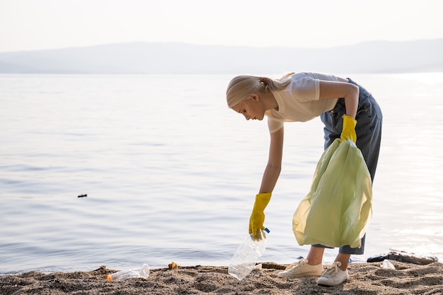 Zdjęcie młoda kobieta umieszcza plastikowe butelki i inne śmieci w plastikowej torbie nad jeziorem. wolontariusz siedzi zgarbiony nad brzegiem jeziora. sobotnie sprzątanie.