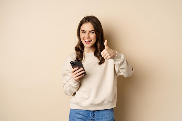 Młoda kobieta uśmiecha się pokazując kciuk w górę podczas korzystania z aplikacji na telefon komórkowy na smartfona, stojąc nad beżową ba...