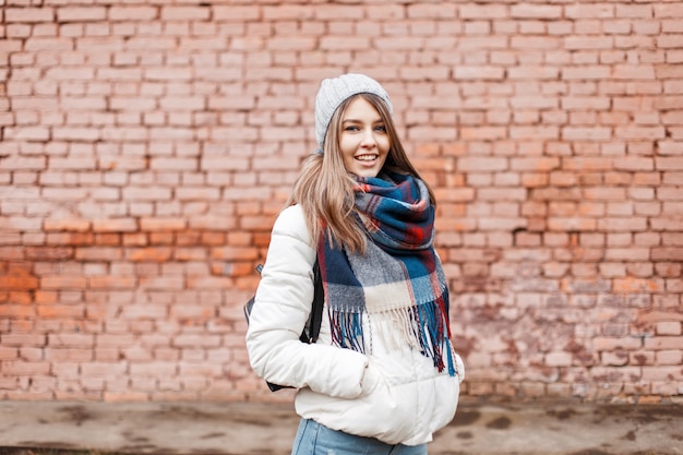 Młoda kobieta uśmiecha się w białą kurtkę i jasny modny szalik blisko ściany
