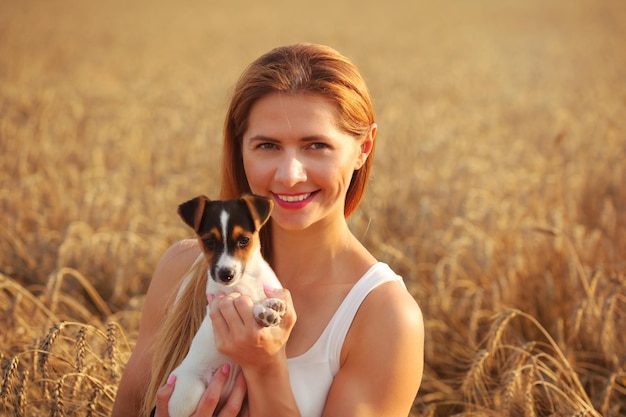 Młoda kobieta uśmiechnięta, trzymając Jack Russell terrier szczeniak, zachód słońca oświetlone pole pszenicy w tle.