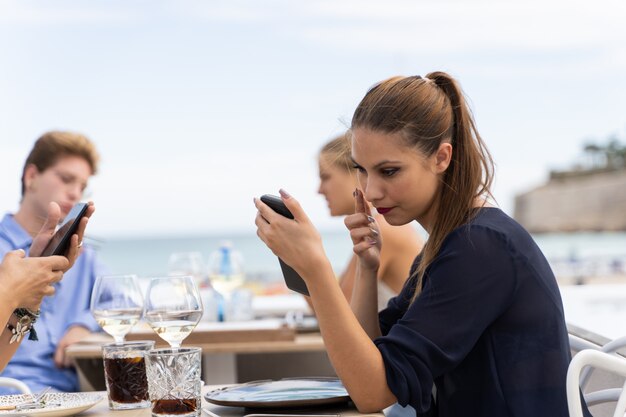 Młoda kobieta używa ekranu smartfona jako lustra do makijażu, siedząc na tarasie restauracji