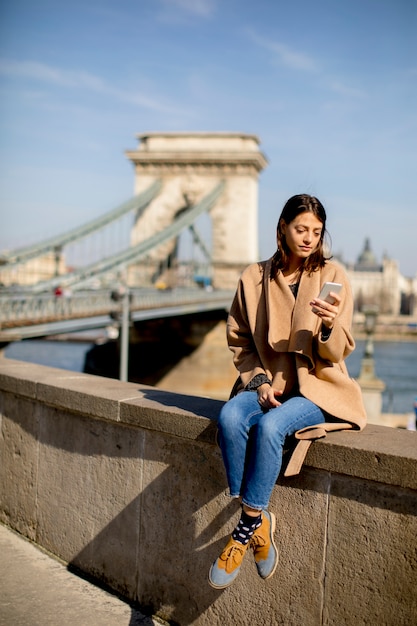 Młoda kobieta używa telefon komórkowego z Łańcuszkowym mostem przy tłem w Budapest