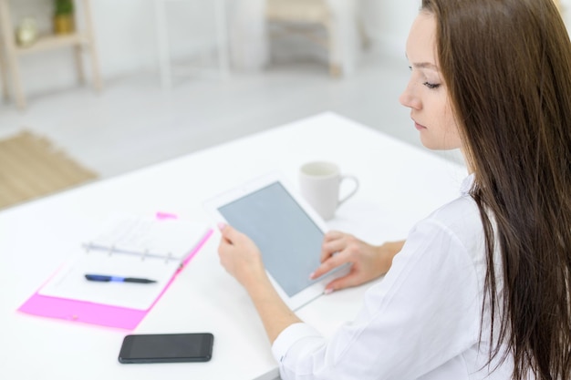 Młoda kobieta w białej koszuli i dżinsach siedzi przy stole z tabletem w dłoniach Kierownik biura wykonuje pracę na nowoczesnym urządzeniu Widok z tyłu