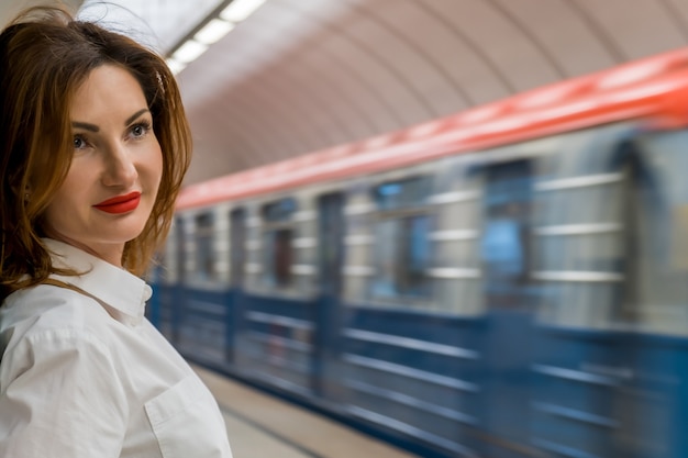 Zdjęcie młoda kobieta w białej koszuli z rudymi włosami i jaskrawoczerwoną szminką stoi na peronie metra na tle przejeżdżającego pociągu. koncepcja transportu publicznego, spotkanie w metrze. miejsce na tekst.