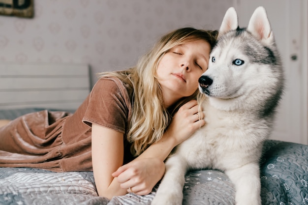 Zdjęcie młoda kobieta w brązowej sukni, leżąc na łóżku w domu z husky szczeniaka.