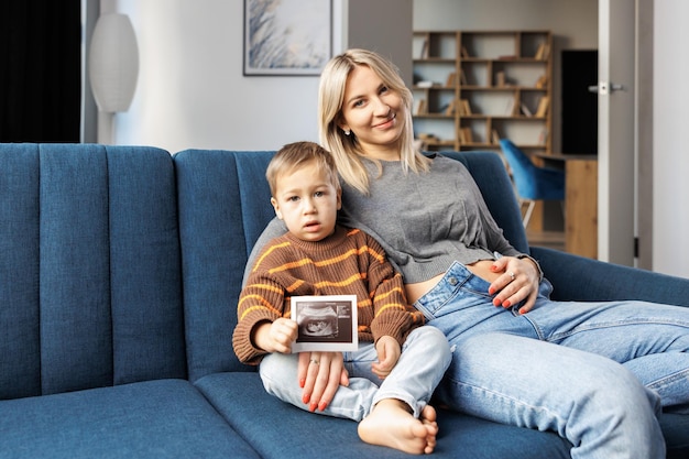 Młoda kobieta w ciąży i jej syn siedzą na kanapie w domu, a chłopiec trzyma w ręku USG Spędzają razem czas, ciesząc się komunikacją, czując kopnięcia nienarodzonego dziecka