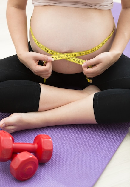 Młoda kobieta w ciąży pomiaru brzucha taśmą po ćwiczeniach na macie fitness