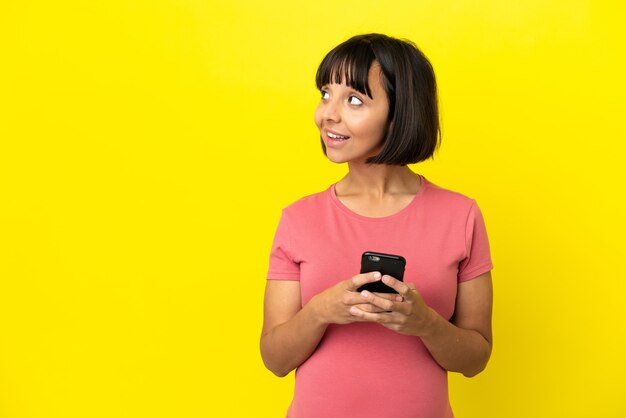 Młoda kobieta w ciąży rasy mieszanej na białym tle na żółtym tle za pomocą telefonu komórkowego i patrząc w górę