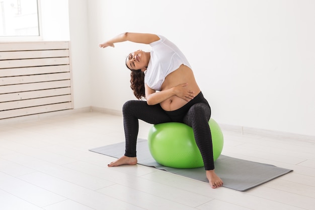 Młoda kobieta w ciąży robi ćwiczenia relaksacyjne przy użyciu piłki fitness, siedząc na macie i trzymając się za brzuch