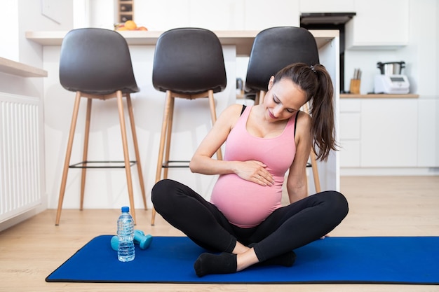 Młoda kobieta w ciąży siedzi na macie do ćwiczeń, dotykając jej brzucha w salonie w swoim domu kaukaskim