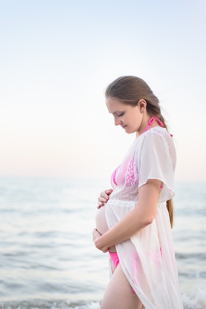 Młoda kobieta w ciąży stoi nad brzegiem morza i ściska brzuch