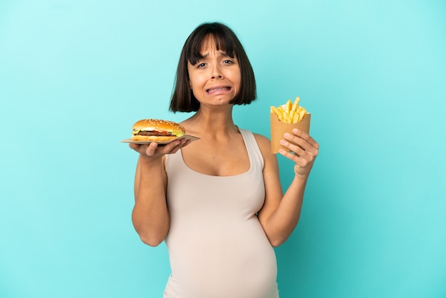 Młoda kobieta w ciąży trzymająca burgera i smażone frytki na odosobnionym niebieskim tle