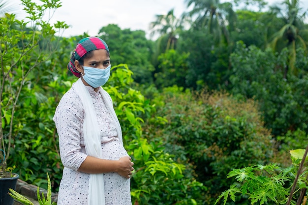Młoda kobieta w ciąży w masce medycznej stoi w zielonej naturze epidemii koronawirusa