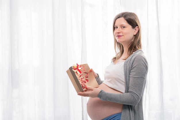 Młoda kobieta w ciąży z dużym brzuchem trzyma prezent owinięty czerwoną wstążką, przyjęcie baby shower z prezentami., prezent i koncepcja szczęśliwego nowego roku.