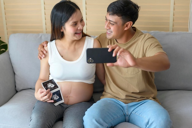 Młoda kobieta w ciąży z mężem w objęciach i rozmowa wideo z rodziną i przyjaciółmi przez smartfona