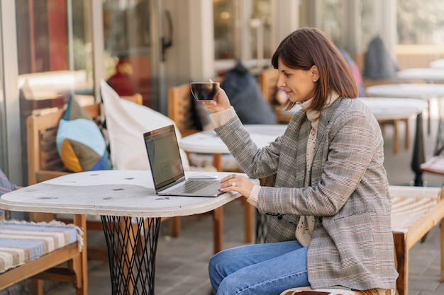 Młoda kobieta w ciepłym swetrze i marynarce pracuje na netbooku w kawiarni na tarasie. Jesienny poranek. Styl uliczny.