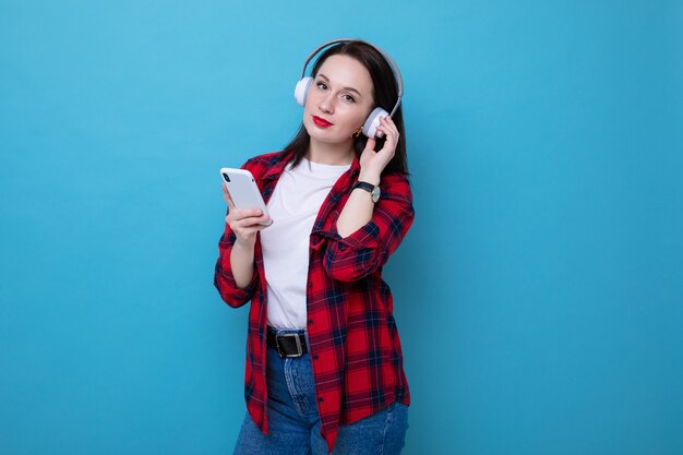 Młoda kobieta w czerwonej koszuli słucha muzyki ze swojego telefonu na niebieskim tle