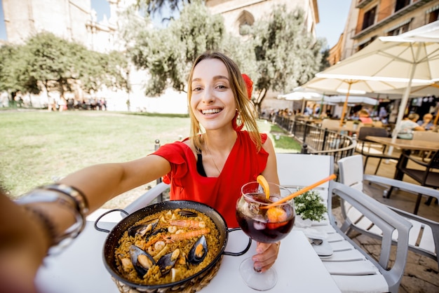 Młoda kobieta w czerwonej sukience robi zdjęcie selfie z morską Paellą, tradycyjnym daniem z ryżu w Walencji i napojem Sangria siedząc na zewnątrz w restauracji w Walencji