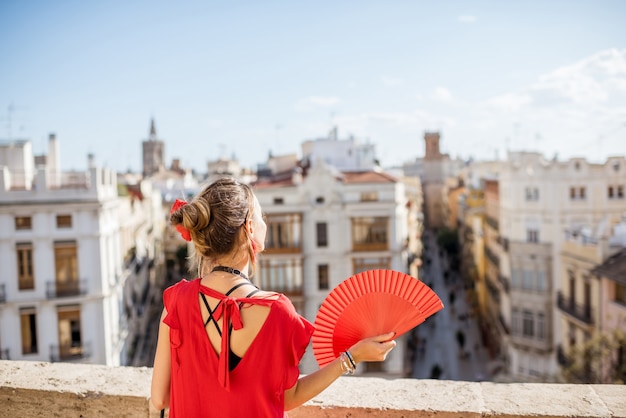 Zdjęcie młoda kobieta w czerwonej sukience z ręcznym wentylatorem i aparatem fotograficznym, ciesząc się pięknym widokiem na miasto walencja podczas słonecznej pogody w hiszpanii