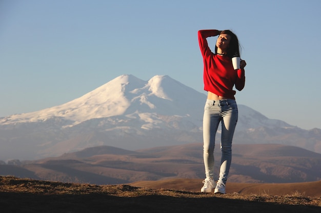 Zdjęcie młoda kobieta w czerwonym swetrze stoi na tle niewyraźnych gór, ciesząc się pierwszymi promieniami słońca z filiżanką herbaty w dłoni
