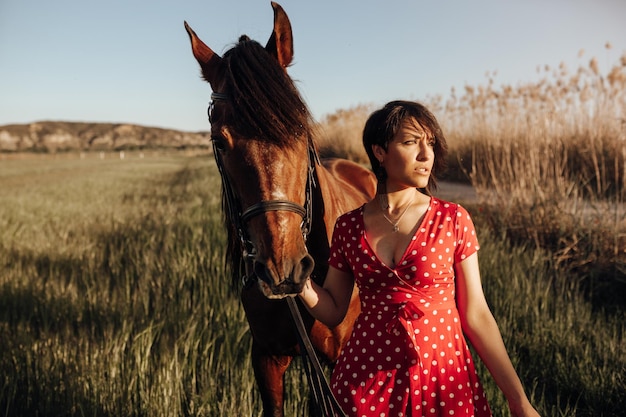 Młoda kobieta w długiej czerwonej sukience z koniem patrząca w stronę, stojąc na podwórku rancza w słoneczny dzień na wsi