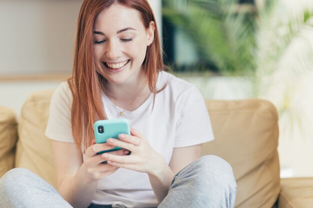 Młoda kobieta w domu odbiera pozytywne emocje wygrywając i dobre wieści podczas czytania telefonu siedząc na kanapie