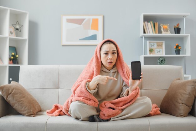 Młoda kobieta w domu ubrania siedzi na kanapie pod kocem w domu wnętrza za pomocą smartfona pokazując na aparat wskazując palcem wskazującym na to patrząc zdezorientowany