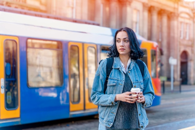 Młoda kobieta w dżinsowej kurtce rozmawia przez telefon i czeka na tramwaj na przystanku