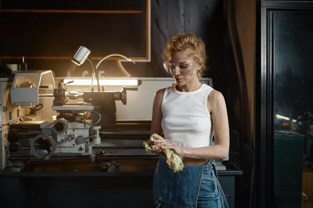Młoda kobieta w dżinsowych kombinezonach i ochronnych okularach zabezpieczających wyciera ręce szmatką po pracy na maszynie obrotowej w warsztacie