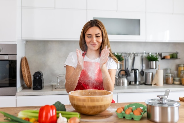 Młoda kobieta w fartuchu przygotowuje ciasto do pieczenia w lekkim wnętrzu kuchni miała pomysł na gotowanie pizzy w domu i wyraz twarzy dmuchający mąkę