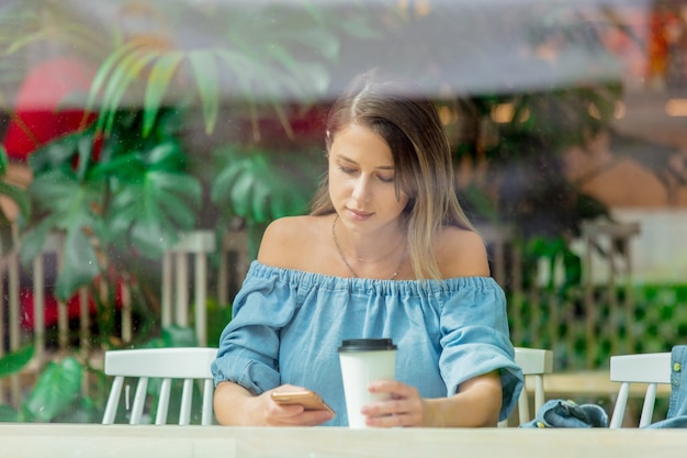 Młoda Kobieta W Kawiarni I Picia Kawy I Korzystania Z Telefonu Komórkowego, Siedząc Przy Oknie