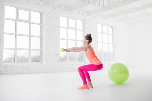 Młoda kobieta w kolorowej odzieży sportowej kuca z hantlami w białej siłowni z piłką fitness na podłodze