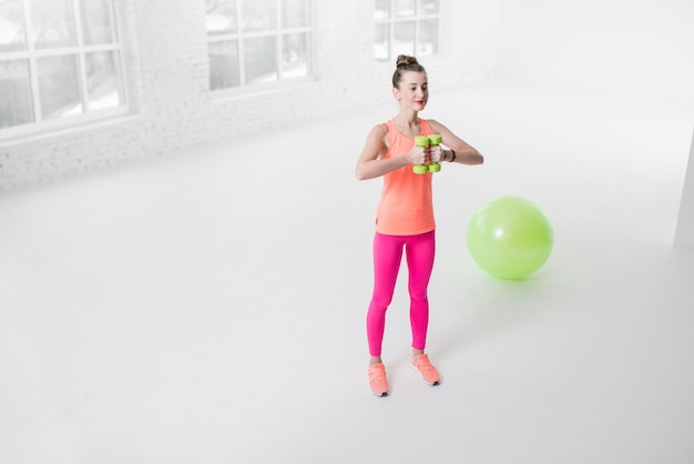 Młoda kobieta w kolorowej odzieży sportowej podnosząca hantle w białym pokoju z zieloną piłką fitness na siłowni