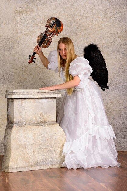 Młoda kobieta w kostiumie anioła trzymająca skrzypce przy ścianie.