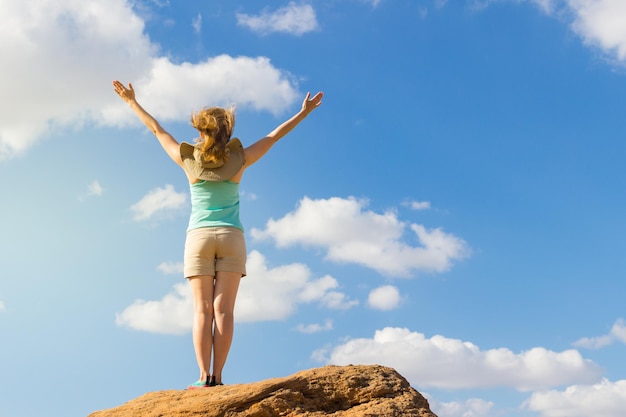 Młoda kobieta w koszuli i szortach z uniesionymi rękami pozostaje na kamieniu. Błękitne niebo z chmurami. Koncepcja sukcesu i wolności.