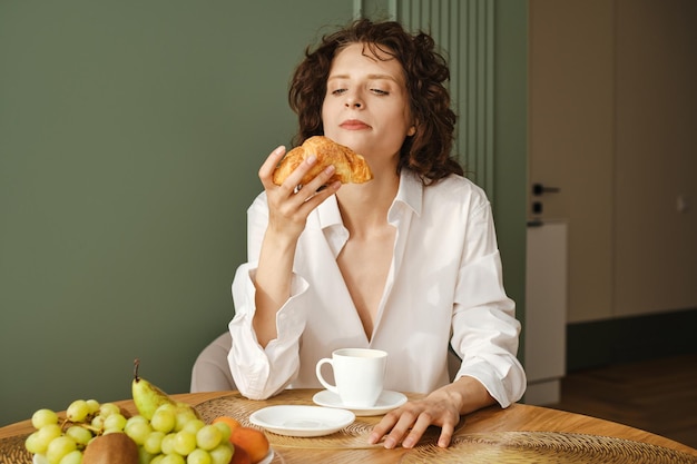 Młoda kobieta w koszuli nad nagim ciałem siedzi przy stole w kuchni i patrzy na croissant, który trzyma w ręku.