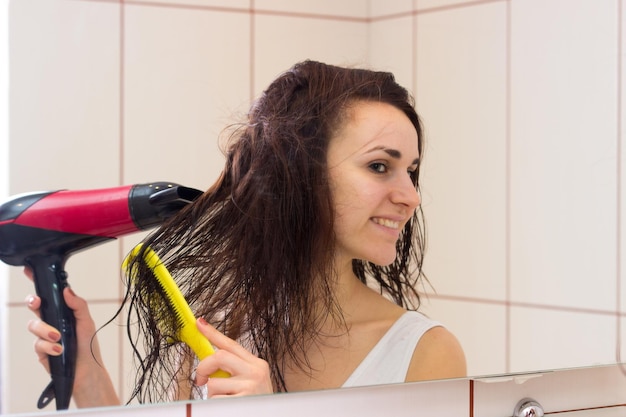 Młoda kobieta w koszuli susząca długie brązowe włosy suszarką do włosów i grzebieniem w łazience