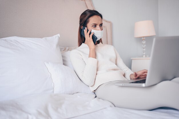 Młoda kobieta w łóżku, mówiąc telefon i pracując na laptopie.