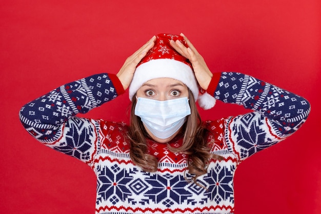 Młoda kobieta w masce medycznej, czapce Świętego Mikołaja i swetrze trzyma głowę z przerażenia lub zaskoczenia.