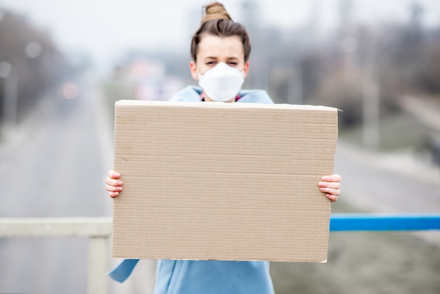 Zdjęcie młoda kobieta w masce ochronnej przed zanieczyszczeniem powietrza trzyma afisz stojący w mieście