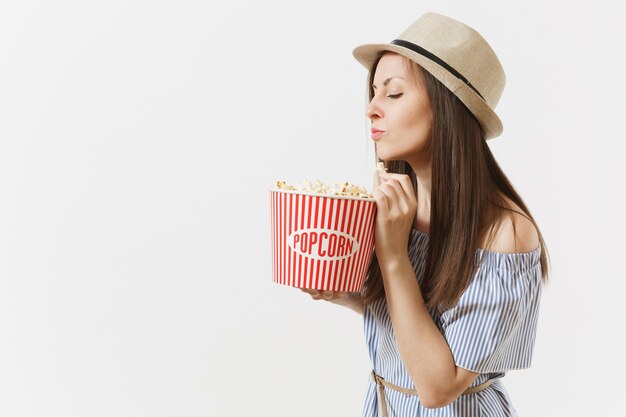 Młoda kobieta w niebieskiej sukience, kapelusz ogląda film trzyma jedzenie popcornu z wiadra na białym tle. Ludzie, szczere emocje w kinie, koncepcja stylu życia. Powierzchnia reklamowa. Skopiuj miejsce.
